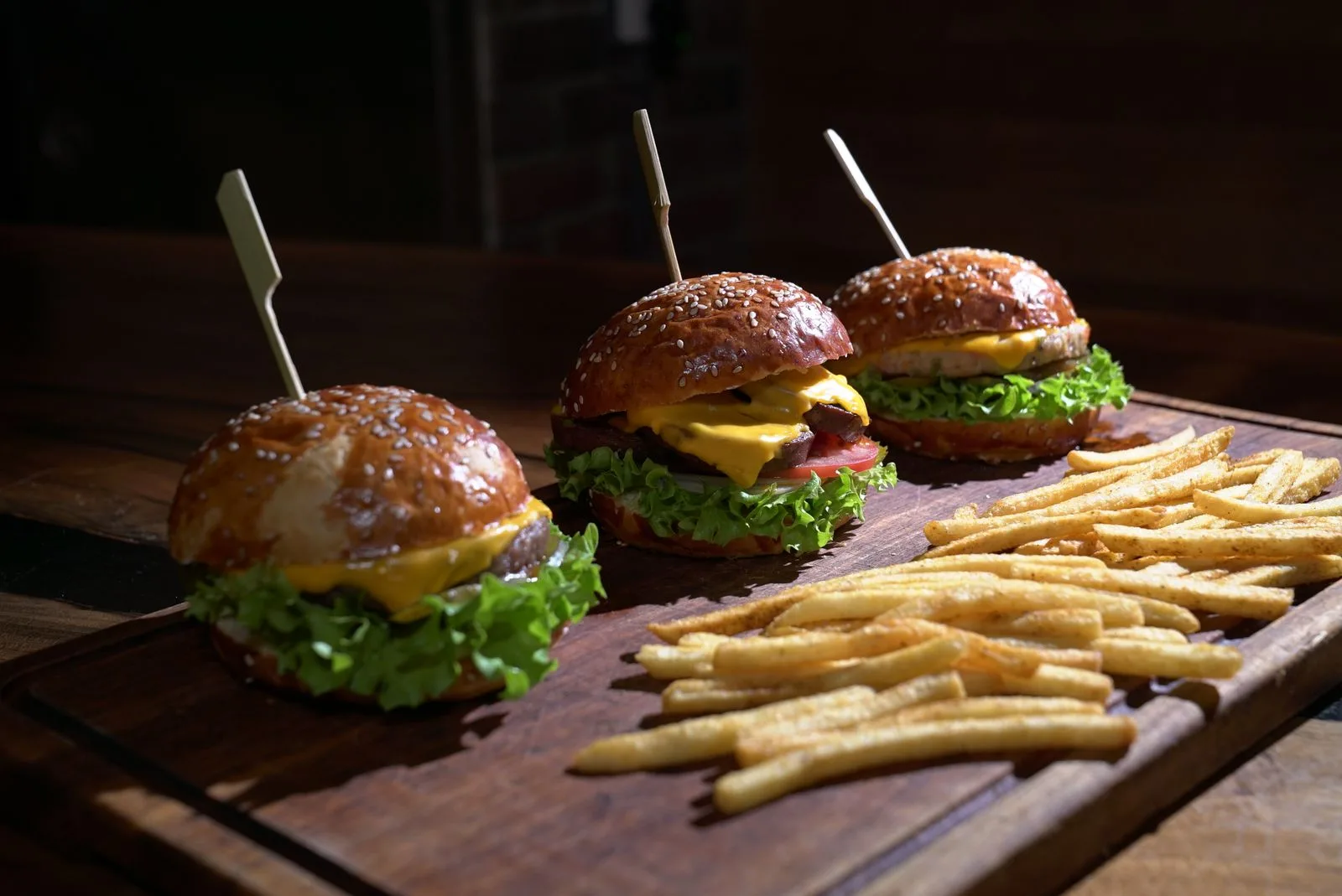 Drei Cheeseburger mit geschmolzenem Käse und Salat neben Pommes frites auf einem dunklen Holzbrett